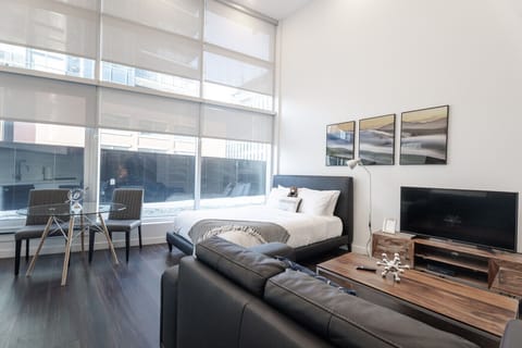 Bright, open-concept studio layout featuring double-height windows, a comfortable king bed, cozy living area with TV, and a glass dining table—offering a seamless blend of modern design and space.