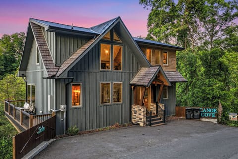 Modern mountain lodge with A-frame architecture sits nestled among tall trees, glowing warmly at dusk. Stone-accented entrance and spacious deck offer peaceful outdoor spaces for your mountain retreat.