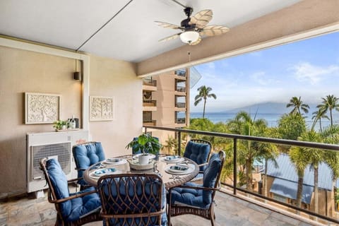 Covered balcony dining area with cushioned chairs and ocean views, framed by palms and tropical décor.