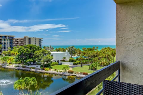 Third-floor balcony with black railing, wicker chair corner, overlooking a pond, palm trees, and ocean horizon.