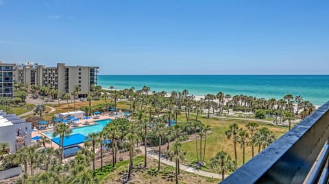 View from the balcony shows a blue pool area with umbrellas, palm trees, sandy beach, and turquoise Gulf waters.