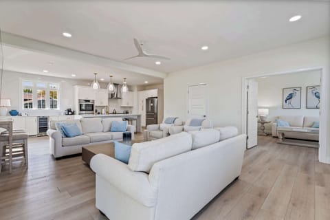 Spacious living room with plush seating, smart TV, electric fireplace, and abundant natural light. Features open-concept layout, high ceilings, coastal décor, and easy access to kitchen and entry—perfect for relaxing or entertaining.