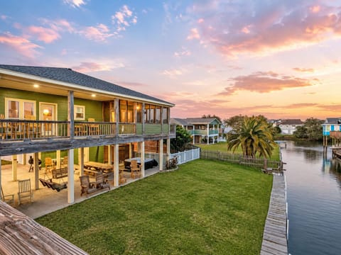 Canal-front Bolivar Peninsula vacation rental with a spacious lawn, wraparound upper deck, covered outdoor living areas and direct waterfront access beneath a colorful sunset sky.
