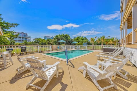 The vacation rental features a private swimming pool surrounded by a spacious deck with lounge chairs, outdoor seating, and a grill—all set within a fenced yard edged by coastal greenery and neighboring beach homes.