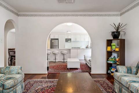 A cozy and well-decorated living space with a stylish archway leading to a modern kitchen area. The living room features patterned sofas, a wooden coffee table, and ornate rugs, while the kitchen has sleek white cabinetry and stainless steel appliances. T