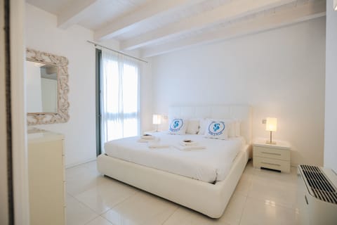Elegant double bedroom in an apartment in Peschiera del Garda, characterized by light colors and a modern, refined design. The room is bright thanks to large windows with light curtains and offers a comfortable king-size bed, bedside tables with lamps, and a large decorative mirror. Perfect for a relaxing stay just a few minutes from the lake.