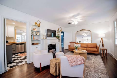 Relax in this stylish living room featuring a warm cognac leather sofa, modern white armchairs, built-in shelving flanking a fireplace with a mounted TV, a large arched floor mirror, dark hardwood floors, and a glimpse into the adjoining kitchen.