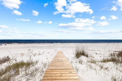 A wooden boardwalk leads across pristine white sand dunes to the expansive ocean shore under a brilliant blue sky.