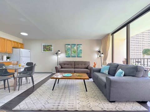 A bright and inviting living space featuring a pull-out sleeper sofa, a couch, tropical artworks, and a connected dining area and kitchen, with a large sliding glass door leading to a balcony.