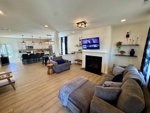 Modern living room with a sectional sofa, fireplace, mounted TV, floating shelves, and open kitchen area.
