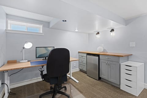 WORKSPACE: Modern setup with a height-adjustable desk, ergonomic chair, built-in counter, mini fridge, and lighting for a comfortable work environment.