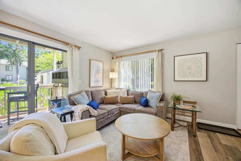 Sun-drenched windows frame this airy living room, where a plush sectional and circular wood table invite slow afternoons. Creamy linens and ocean-blue accents harmonize with the golden light spilling in from the private balcony.