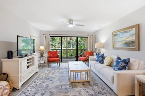 Vibrant coral armchairs and nautical pillows add pops of color to this airy living room. Sliding glass doors frame a lush green view, inviting the soft afternoon glow to settle over the plush rug and clean white finishes.