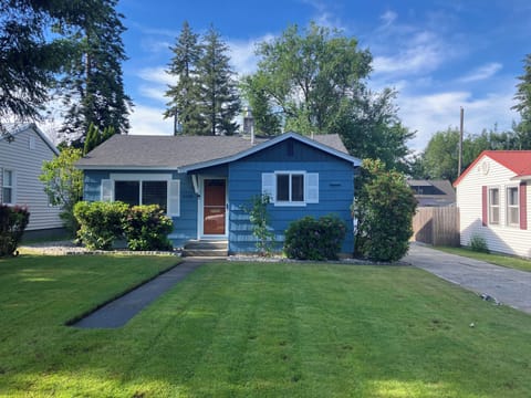 A charming blue bungalow sits on a quiet residential street, featuring mature landscaping and a tree-lined setting. A convenient parking area completes this welcoming neighborhood retreat.