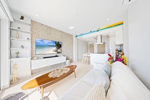 Modern living room with a white sofa and a surfboard-shaped coffee table. The TV is mounted on a textured mosaic wall, and built-in white shelves are on the left. The kitchen is visible in the background. A long, narrow painting of a beach scene is mounte