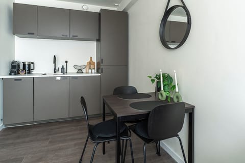 Modern kitchenette with sleek grey cabinets, sink, coffee machine, kettle, and a cozy dining table for two in a clean, stylish space.