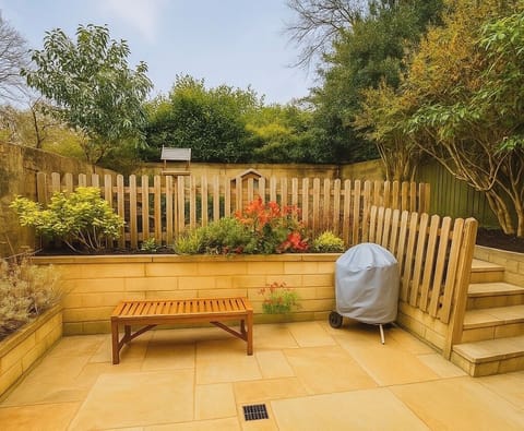 Tranquil patio with a wooden bench, BBQ grill, and a raised garden behind a picket fence. Perfect for outdoor relaxation or dining in a charming setting with natural stone paving.