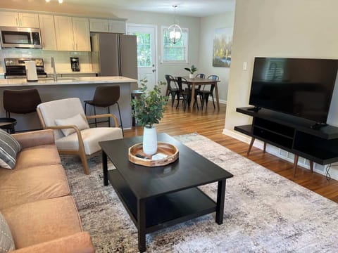 Stylish open-concept living space featuring a cozy sofa, modern coffee table, and plush area rug. The living room flows into a sleek kitchen with bar seating and a bright dining area, creating a warm, welcoming setting for relaxing or entertaining.