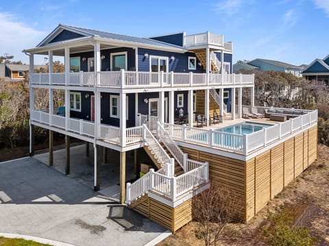 Large blue beach house on pilings with wraparound white rail decks, rooftop lookout, private pool deck with seating, outdoor dining area, stairs, covered parking, and bright coastal sky backdrop.