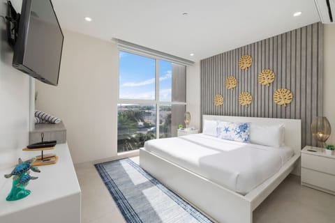 Stylish bedroom with a white platform bed and a textured gray accent wall decorated with gold leaf-shaped art. A large window offers a bright view, and the room has a blue and white rug.