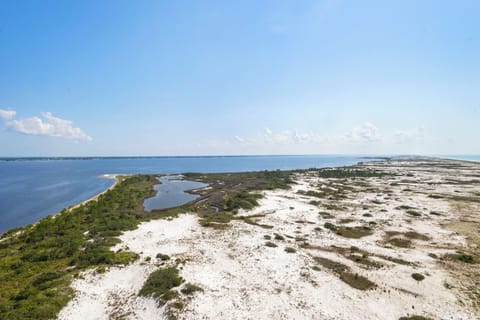 Aerial view captures the coastal peninsula with sandy beaches and natural vegetation bordering calm blue waters under a bright sky.