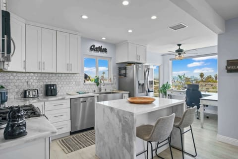 This modern kitchen features sleek white cabinetry, a marble island with bar stools, stainless steel appliances, and a bright open layout that flows into a cozy home office with scenic window views.
