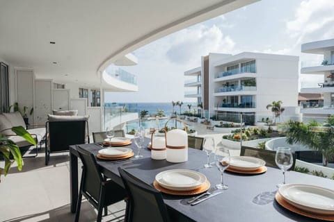 Luxurious oceanfront balcony with a modern dining set and a view of a sleek white resort and the sparkling blue sea.