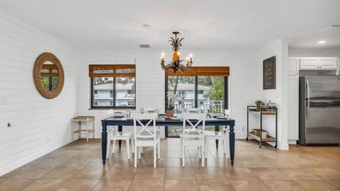 Bright and coastal dining area with a table set for eight, offering views of the charming homes and condos along the street. Features include dinner service for eight, a bar cart ready for cocktail hour, and access to the kitchen for seamless entertainment