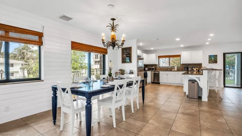 Bright and coastal dining area with a table set for eight, offering views of the charming homes and condos along the street. Features include dinner service for eight, a bar cart ready for cocktail hour, and access to the kitchen for seamless entertainment