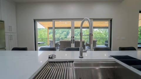 Modern kitchen island with a deep stainless steel sink and spring-neck faucet, offering a stunning view through sliding glass doors to a covered patio and lush, tree-filled valley beyond.