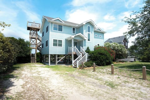 Charming coastal vacation home features elevated construction with stilts, private observation tower, spacious parking, and easy beach access surrounded by lush landscaping and neighboring properties.