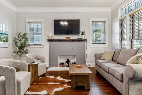 The bright living room pairs comfortable seating with a decorative fireplace, large smart TV, original windows, and warm hardwood floors.