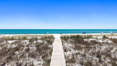 A wooden boardwalk leads through natural dunes to pristine white sand and sparkling turquoise waters, where beachgoers relax under umbrellas on this beautiful coastal setting.