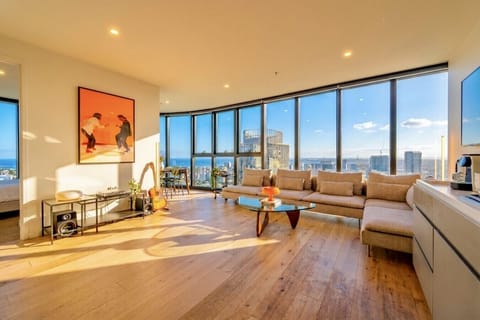 Open-plan living area with floor-to-ceiling curved windows framing skyline and ocean views. Warm timber floors, a large sectional sofa, vinyl turntable, and curated art create a relaxed, design-led retreat.