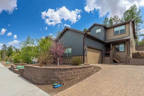 Modern mountain home with attached garage, landscaped beds, and paver driveway set among tall pines under a bright blue sky.