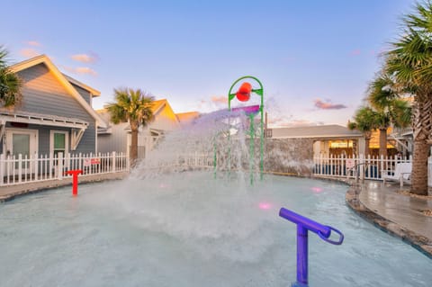Kids' splash zone features colorful water play equipment surrounded by palm trees and charming coastal cottages at twilight.
