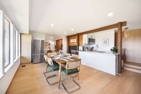 A sleek kitchen with white cabinets flows into a dining area with green-cushioned chairs and wood beams.
