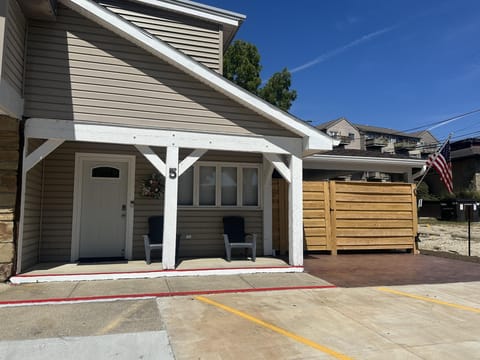 Covered entry porch with beige siding, white trim, two gray chairs, and a wooden privacy fence beside parking spaces.