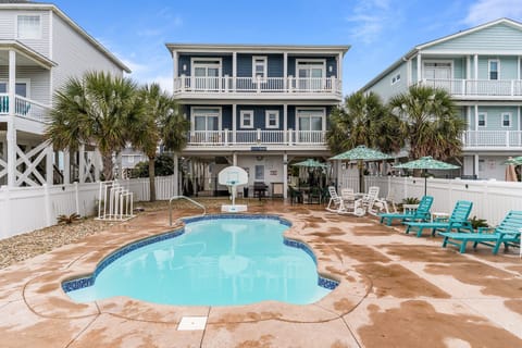 Oak Island NC vacation rental with a private fenced backyard featuring an in-ground pool, wet concrete deck, teal lounge chairs, patio seating, umbrellas, palm trees, outdoor basketball hoop, and elevated beach house with balconies near the ocean.