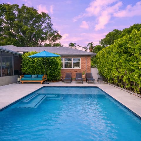 Inviting backyard showcases a sparkling blue swimming pool surrounded by a paved area and lush green landscaping under a clear sky.
