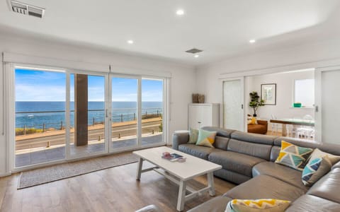 A modern, stylish living room with a large sectional sofa adorned with colorful cushions. The room features expansive sliding glass doors that offer a stunning view of the ocean, creating a serene and inviting atmosphere. The interior is bright and airy, 