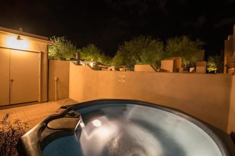 A hot tub is illuminated at night, surrounded by a low wall and plants, with a door visible in the background.