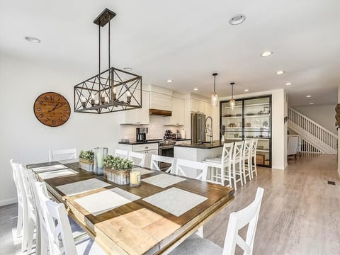 The spacious dining area showcases a beautiful wooden table with white cross-back chairs, seamlessly connecting to a modern kitchen equipped with stainless steel appliances, a Keurig, and stylish pendant lighting.