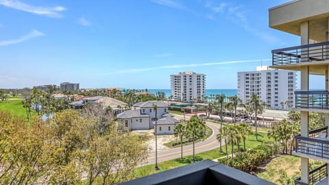 Corner balcony railing frames palm trees, manicured golf course, and distant white condos against a blue sky.