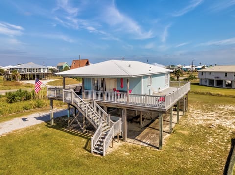 Elevated beach house with wraparound deck and stairs, situated in a coastal neighborhood with other vacation homes nearby.