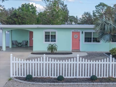 Charming mint-green home with a coral front door, large windows, and a white picket fence. The front entry is neat and welcoming with simple landscaping and a covered seating nook.