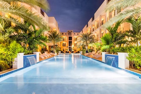 A tranquil resort pool surrounded by lush palm trees, with soft lighting illuminating the white buildings. The clear water reflects the warm lights, creating a serene, welcoming atmosphere at night.