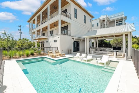 Modern coastal architecture featuring multiple covered decks, private pool with sun shelf, and outdoor lounge pavilion set among lush tropical landscaping under blue skies.