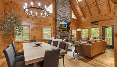 Beautiful and cozy interior of our modern cabin. The space features an open-concept living and dining area with an abundance of natural wood. The living area boasts a grand stone fireplace that extends up to the vaulted ceiling.