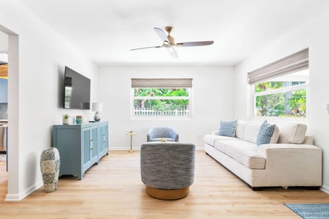 Bright living room with wall-mounted TV, teal console, linen sofa, and garden-facing windows — easygoing coastal style throughout